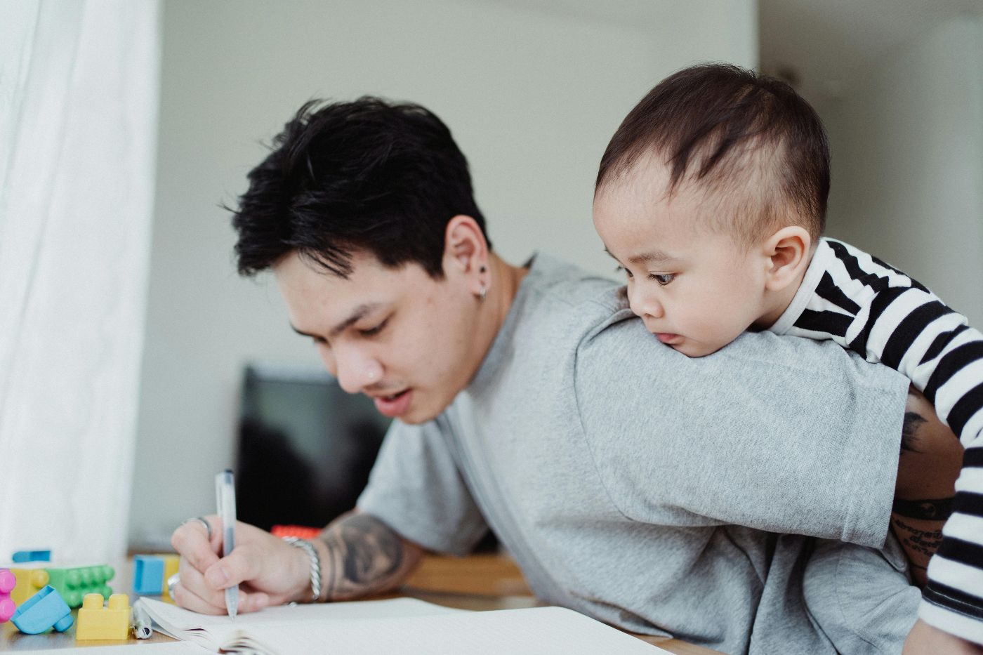 Kleinkind sitzt auf Schultern seines Vaters, während Vater am Schreibtisch einen Arbeitsauftrag erledigt. Foto symbolisiert Vereinbarkeitsthemen.