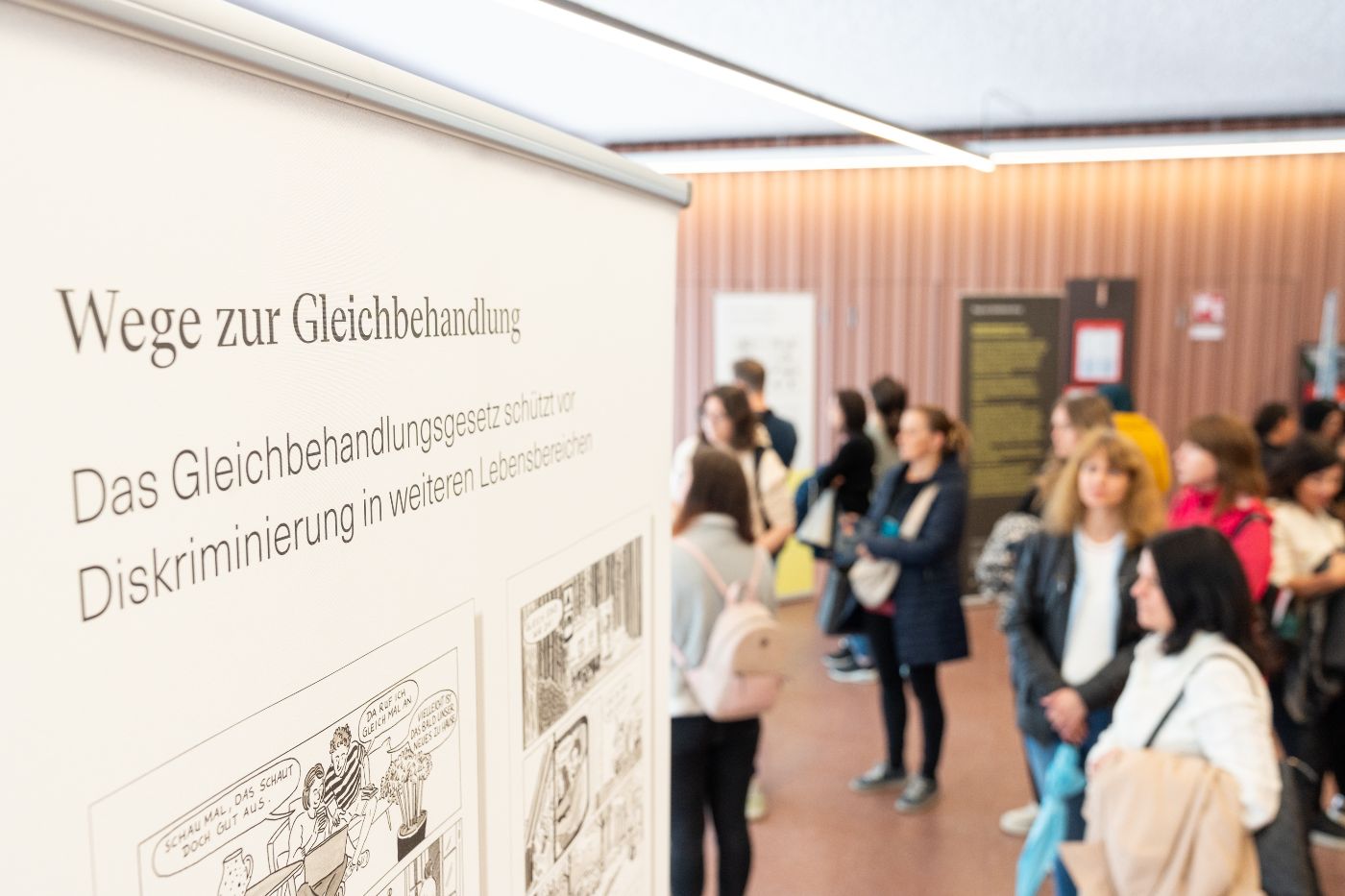 Ein Teil der Wanderausstellung. In der linken Bildhälfte im Vordergrund sieht man den oberen Teil eines Roll-Ups, dort steht "Wege zur Gleichbehandlung. Das Gleichbehandlungsgesetz schützt vor Diskriminierung in weiteren Lebensbereichen". Auf der rechten Bildhälfte sind im Hintergrund Besucher:innen der Ausstellung zu sehen